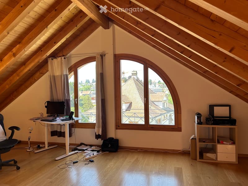 Bright attic room with wooden slanted ceilings, large arched windows, and modern furniture including a standing desk, ergonomic chair, laptop, and printer.