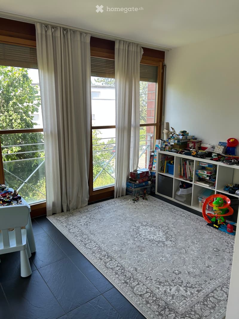 Bright room with large windows and balcony access, featuring a patterned rug, a small white chair, a shelf filled with books and toys, and a small table with toys on it.