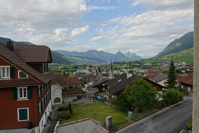 View from a rooftop showing a town with houses, a church spire, and distant mountains under a partly cloudy sky.