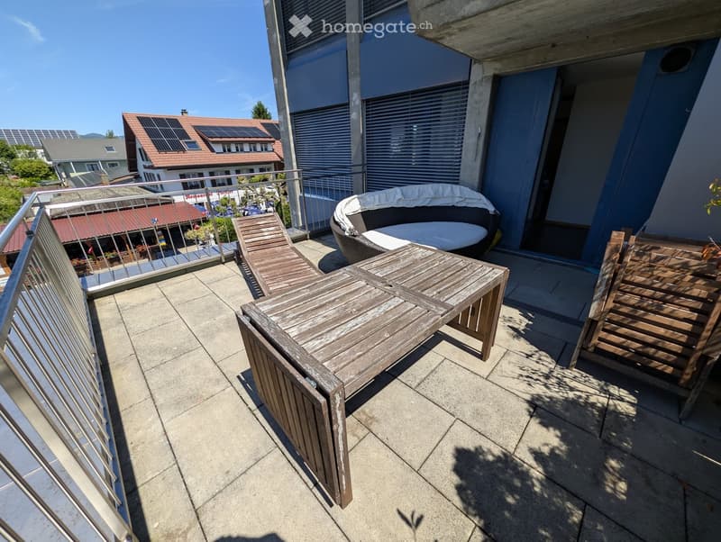 Balcony with wooden table, chairs, and a sofa, overlooking a residential area with solar panels on neighboring roofs.