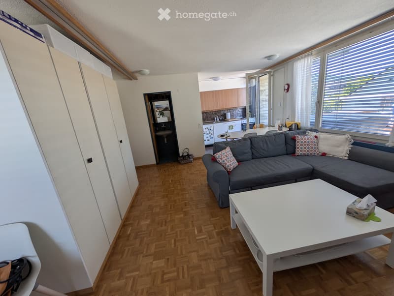 Bright living room with grey sectional sofa, white coffee table, and large windows with blinds. Adjacent kitchen area visible with cabinets, refrigerator, and dining table.