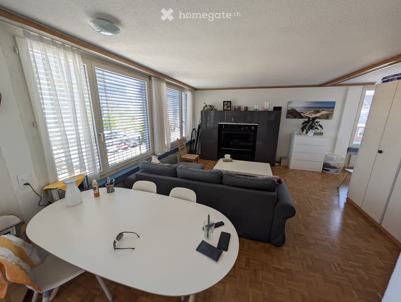A modern living room with a grey sofa, coffee table, fireplace, and large windows with blinds. The room features a round dining table with chairs, a small cabinet, and various decorative items.
