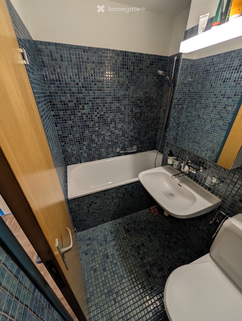 Compact bathroom with blue mosaic tiles, featuring a bathtub, sink, toilet, and showerhead.