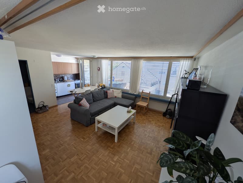 Living room with wooden flooring, grey sectional sofa, white coffee table, small wooden chair, dining table with chairs, and a kitchen area with cabinets and appliances.
