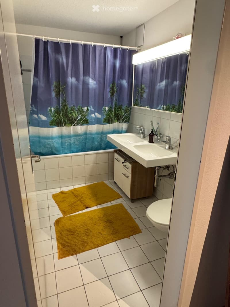 Bathroom with white tiles, double sink, toilet, mirror, and a shower with a tropical-themed curtain. Two yellow rugs are placed on the floor.