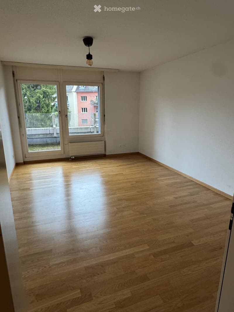 Empty room with wooden flooring, white walls, and a large window with a view of the outside. There is a radiator below the window and a ceiling light fixture.