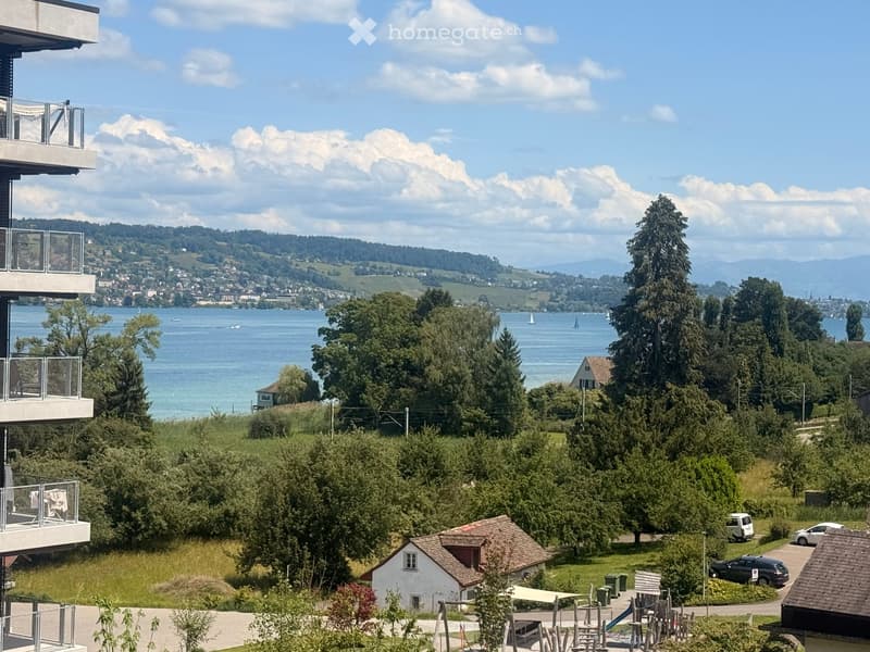 Balcony overlooking a scenic lake with sailboats, surrounded by trees and distant hills under a partly cloudy sky.