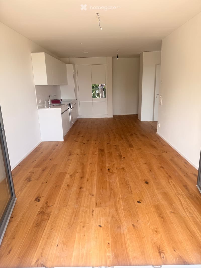 Empty room with light wooden flooring and white walls, featuring a small kitchen area with cabinets and a sink.