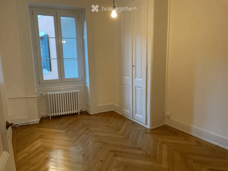 Empty room with wooden herringbone flooring, white walls, a single window with shutters, a radiator, and a closed white door.