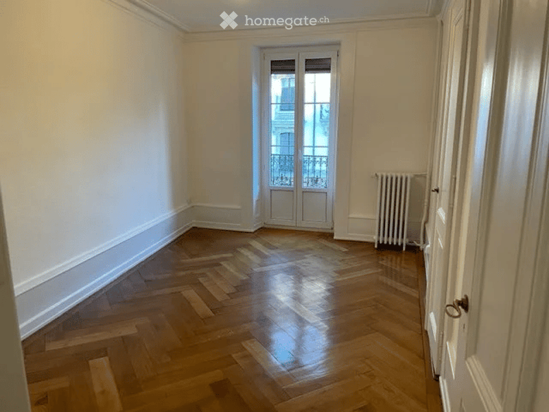 Empty room with wooden parquet flooring, white walls, and a radiator. A door leads to a balcony with a metal railing visible through the glass.