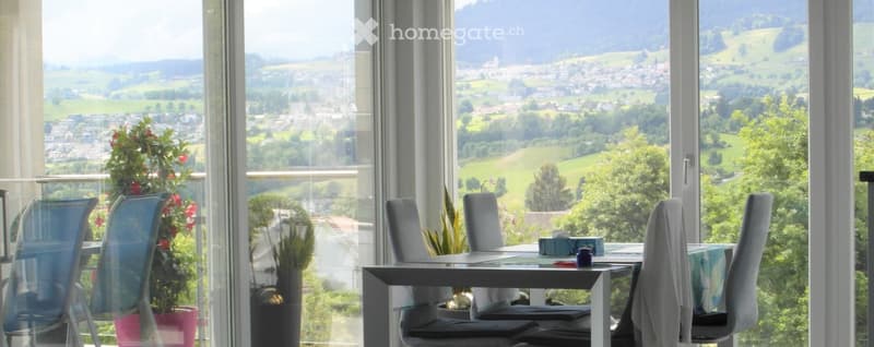 Balcony with a scenic view of a town and countryside, featuring a modern dining table, chairs, and potted plants.