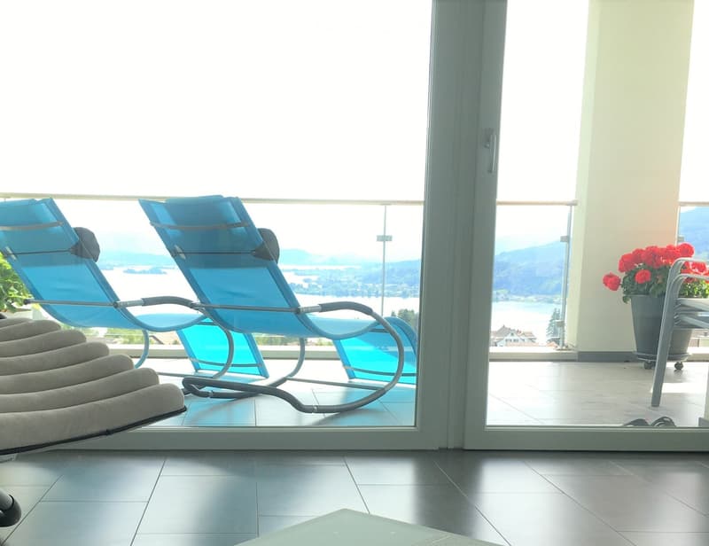 Balcony with lounge chairs, a small table, and a flower pot, offering a scenic view of a lake and mountains.