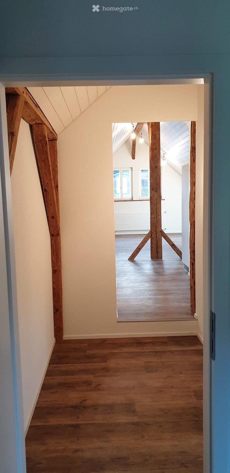 Corridor / Hallway. A narrow hallway with wooden flooring and white walls, leading to an open space with wooden beams and large windows.