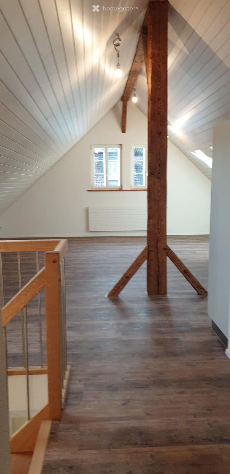 Unfurnished Room. An empty attic space with wooden floors, exposed wooden beams, and a slanted white ceiling. There are two windows at the far end and a wooden staircase railing visible on the left side.