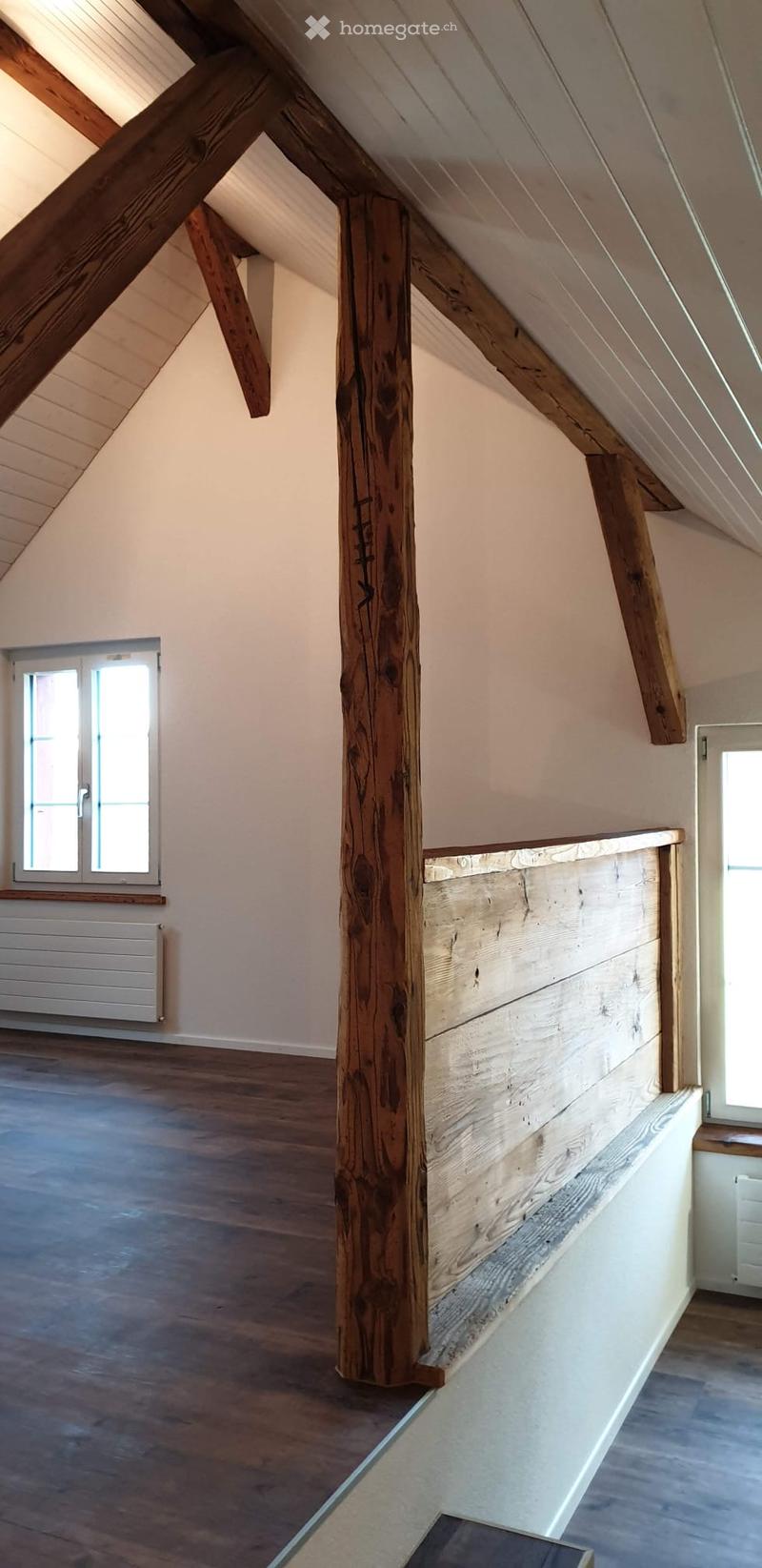 Unfurnished Room. An empty attic space with wooden beams, wooden floor, and large windows allowing natural light. The room features exposed wooden beams and a wooden railing along the edge of the loft area.