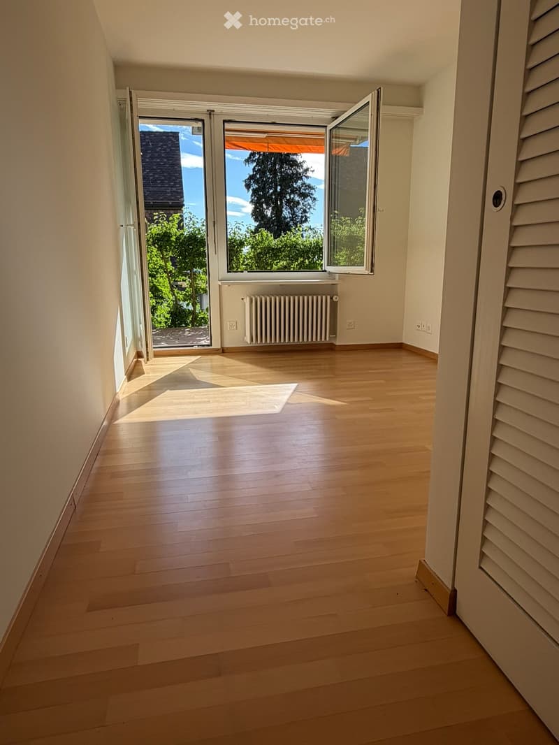 Empty room with wooden flooring, large windows, radiator, and natural light streaming in.