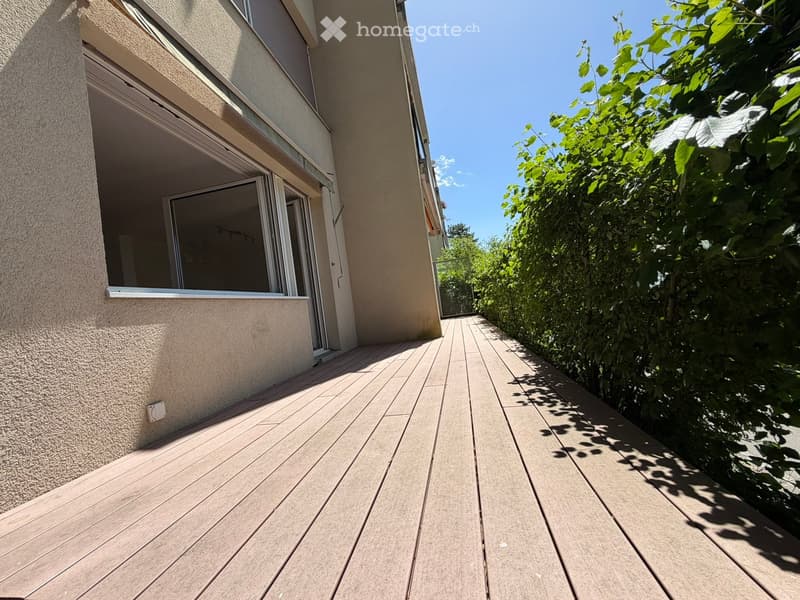 Wooden deck leading to a building with large windows, surrounded by lush greenery.