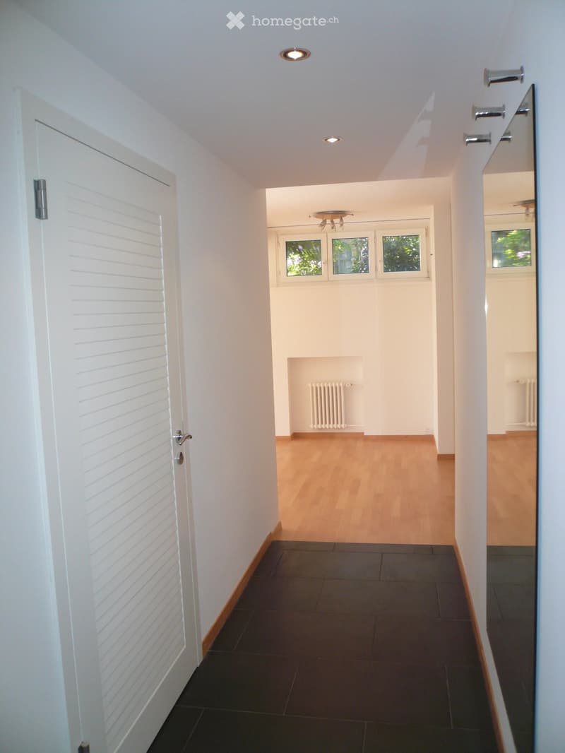 Bright hallway with white walls, wooden flooring, and recessed lighting. Features a white louvered door on the left and a radiator in the adjacent room.