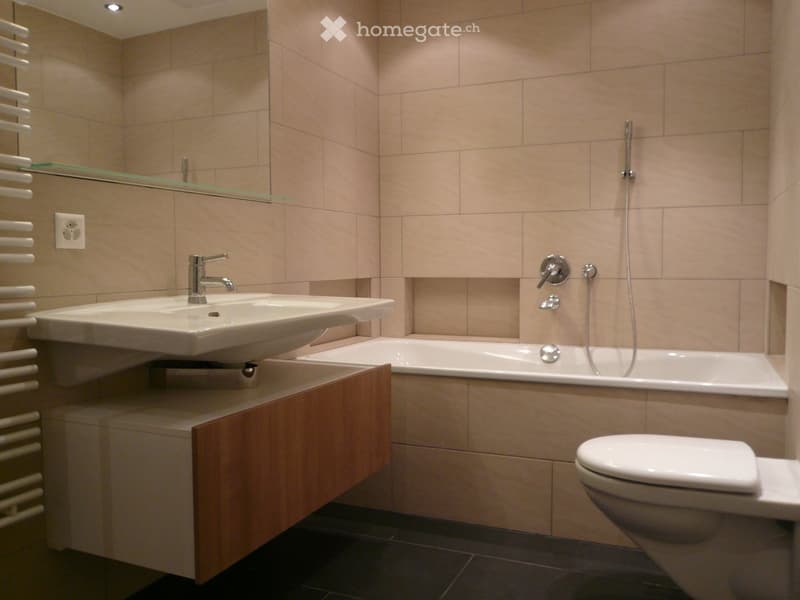 Modern bathroom with beige tiles, a double sink, a bathtub, a toilet, a mirror, and a radiator.