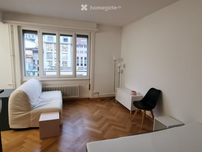 A small, well-lit room with wooden flooring, large windows, a sofa, a chair, a small table, and a lamp.