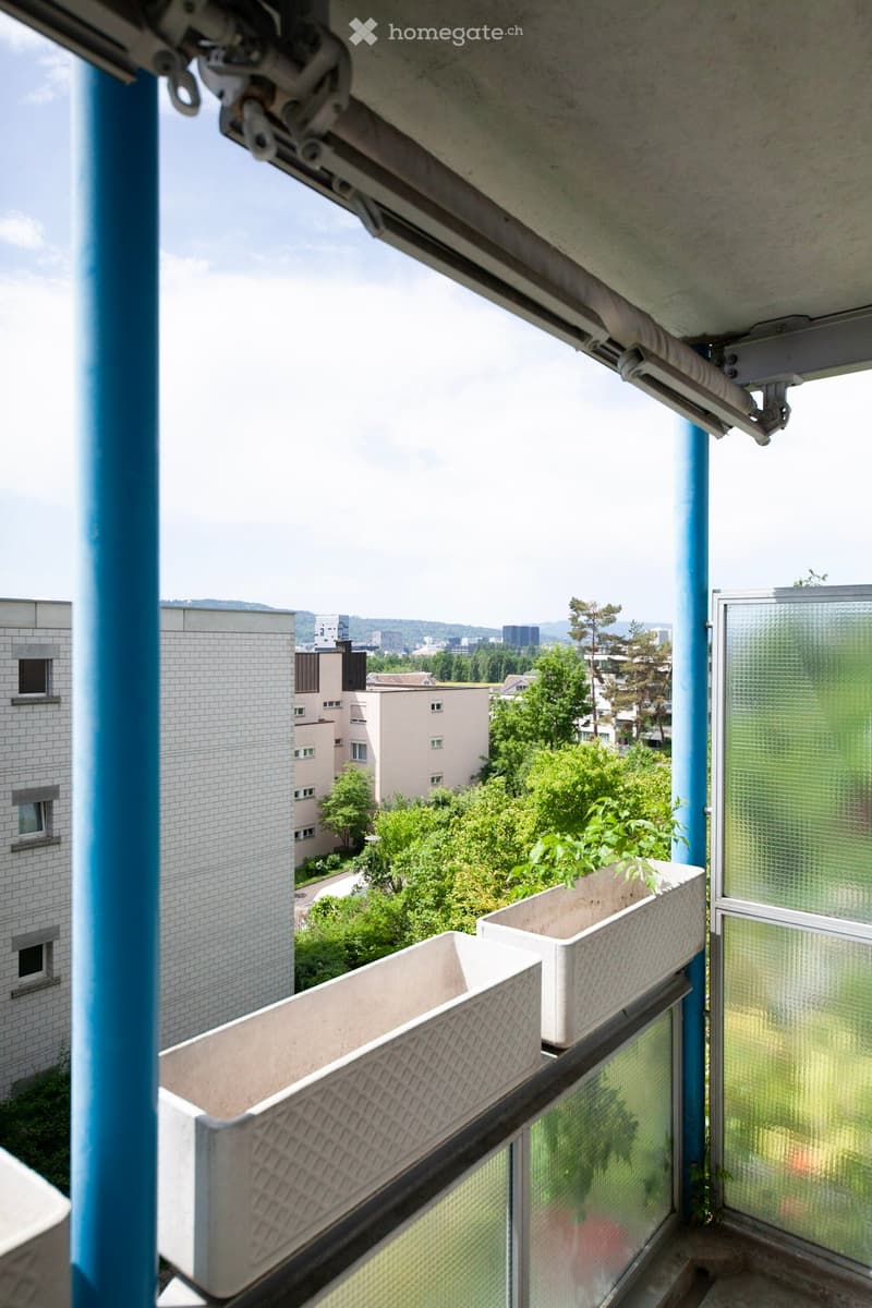 Balcony with two white planters, overlooking a cityscape with buildings and greenery.