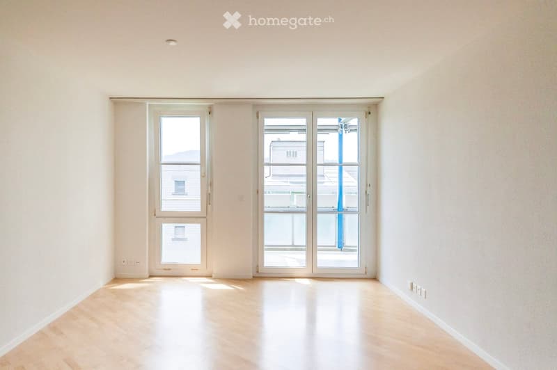 Empty room with light wooden flooring and white walls, featuring large windows and a sliding glass door leading to an outdoor area.
