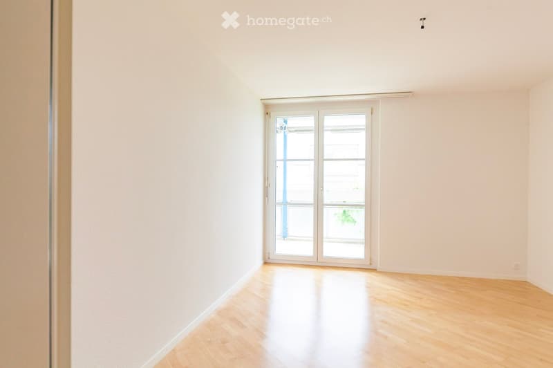 Empty room with light wooden flooring and white walls, featuring a large sliding glass door leading to a balcony.