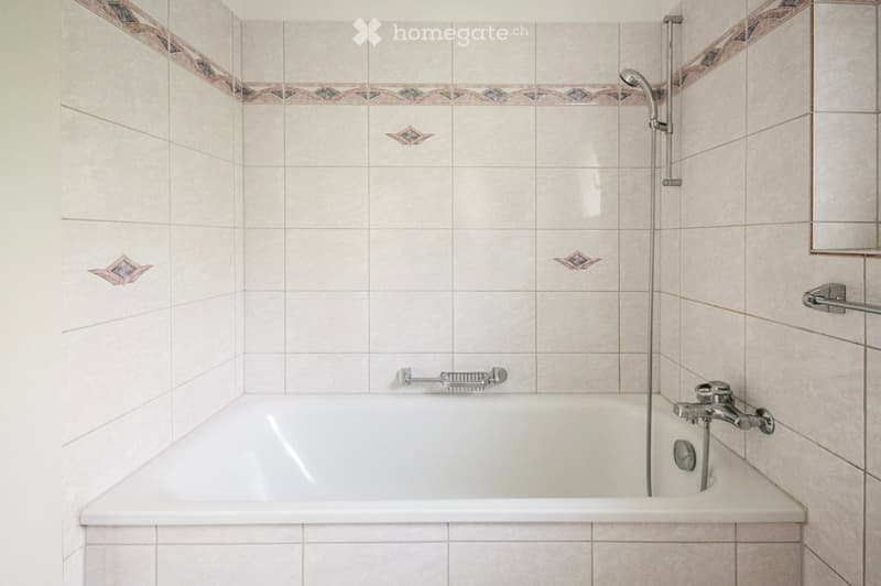 Bathtub with modern faucet and showerhead, tiled walls with decorative border, handrail for safety.