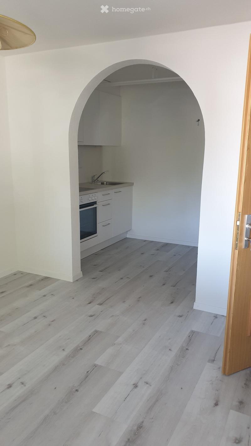 Empty room with light wooden flooring, white walls, and a small kitchen area with cabinets, sink, and oven visible in the background.