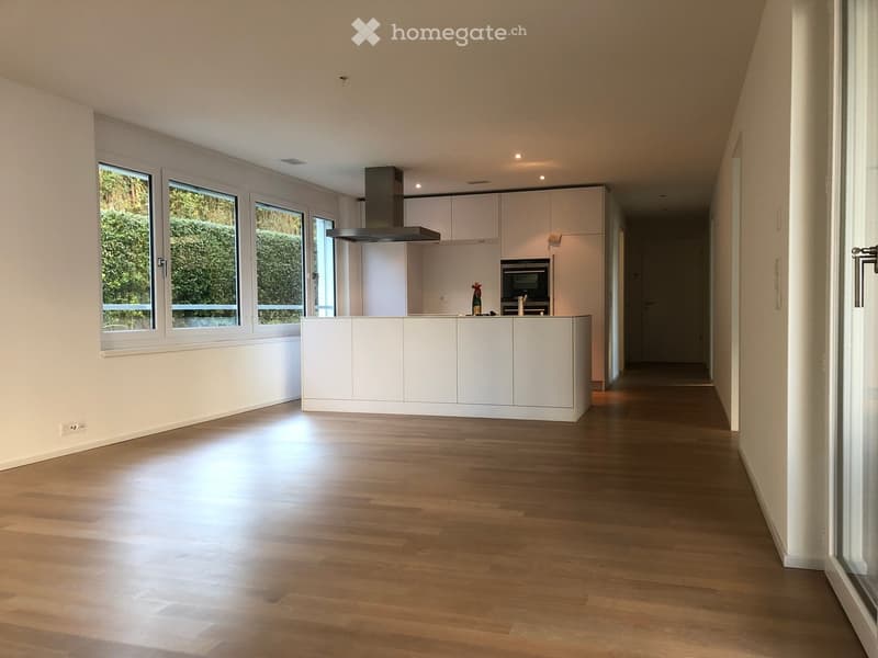 Bright hallway with wooden flooring, white walls, and large windows on one side, leading to a modern kitchen area with white cabinets and stainless steel appliances.