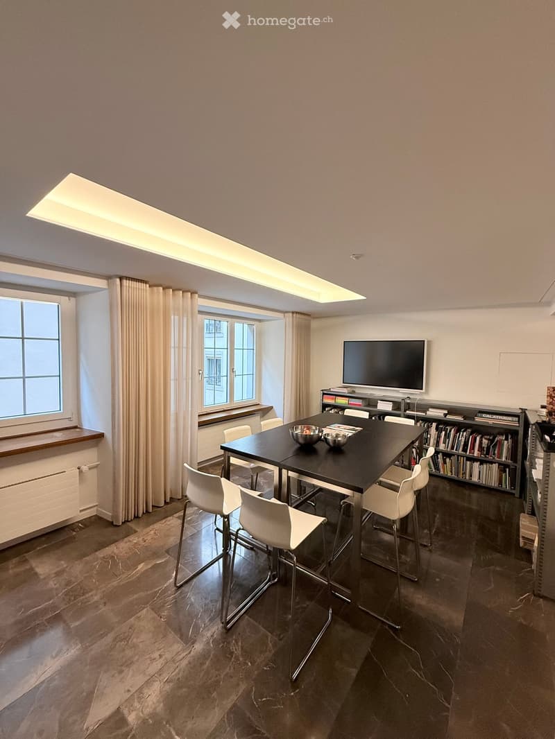 Modern dining area with a black table, white chairs, a large screen, and a bookshelf filled with books.