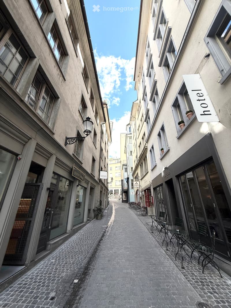 Narrow cobblestone alleyway flanked by multi-story buildings with shops and cafes, featuring outdoor seating and signage for a hotel.