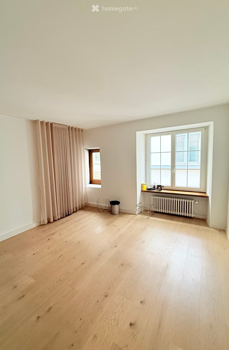 Empty room with light wooden flooring, white walls, and large windows with curtains. Minimal decor includes a radiator, a small shelf, and a trash bin.