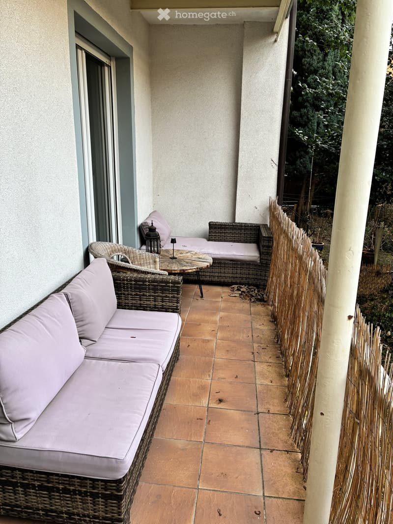 Balcony with wicker furniture, including a sofa, a table, and a chair, with a view of greenery outside.