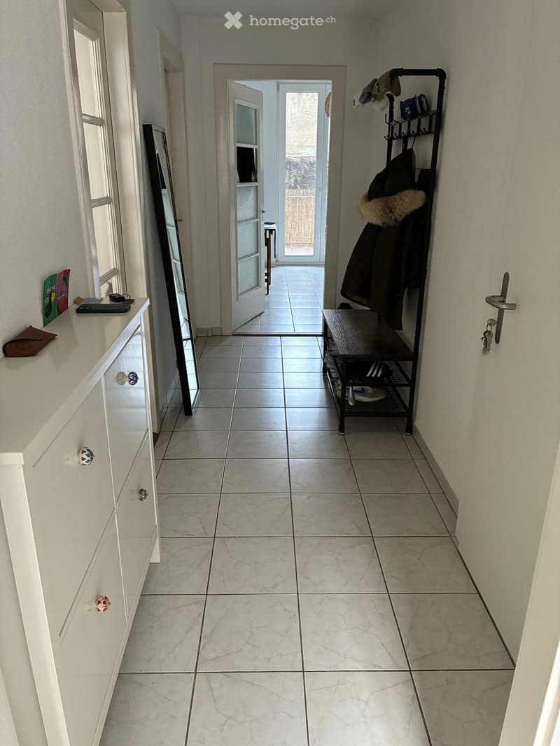 Clean, tiled hallway with white walls, a coat rack, a small table, and a mirror. The hallway leads to an open door showing a view of the outdoor area.