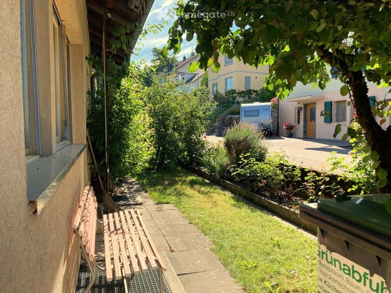 A small garden area with a bench, a tree, and a trash bin. The garden is surrounded by a concrete pathway and has a grassy lawn. There are plants and flowers in the garden, and a building with a door and a window is on the right side. A camper van is park