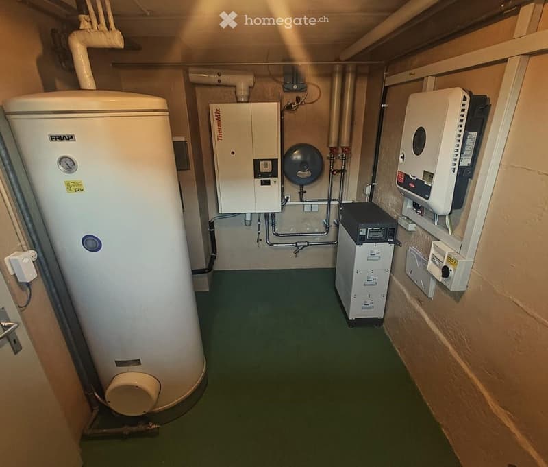 A room with a large white water heater, a white ThermiMix unit, and a white inverter unit mounted on the wall. There are pipes and wires connected to the units. The room has a green floor and beige walls.