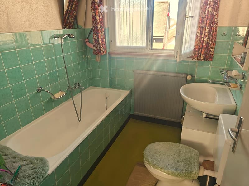 Bathroom with green tiles, bathtub, shower head, sink, toilet, radiator, towel, soap dish, brush, mirror, window, and curtain.