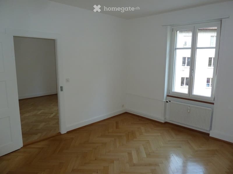 Empty room with light-colored wooden herringbone flooring, white walls, and a large window with white frames and a radiator below it. The room has a door leading to another space.
