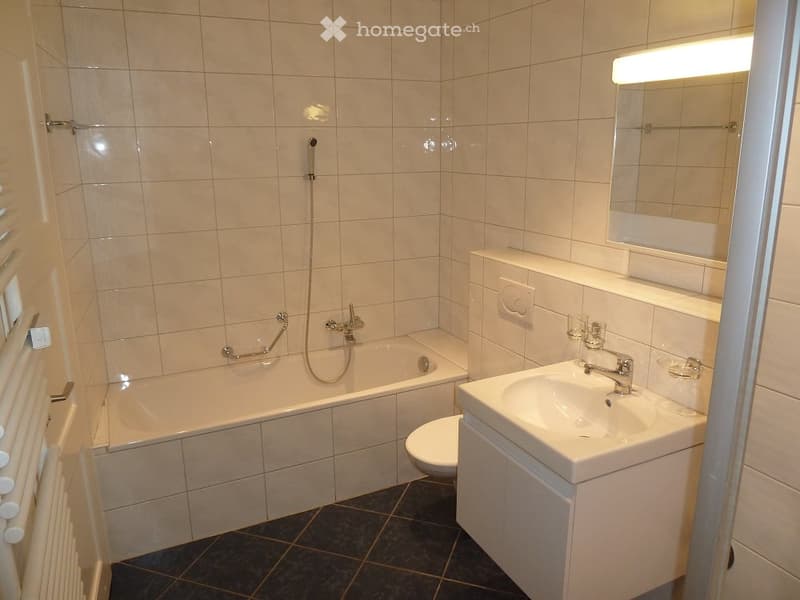 Modern bathroom with a bathtub, toilet, sink, mirror, and showerhead. The walls and bathtub are tiled in light beige, and the floor is dark tiled.