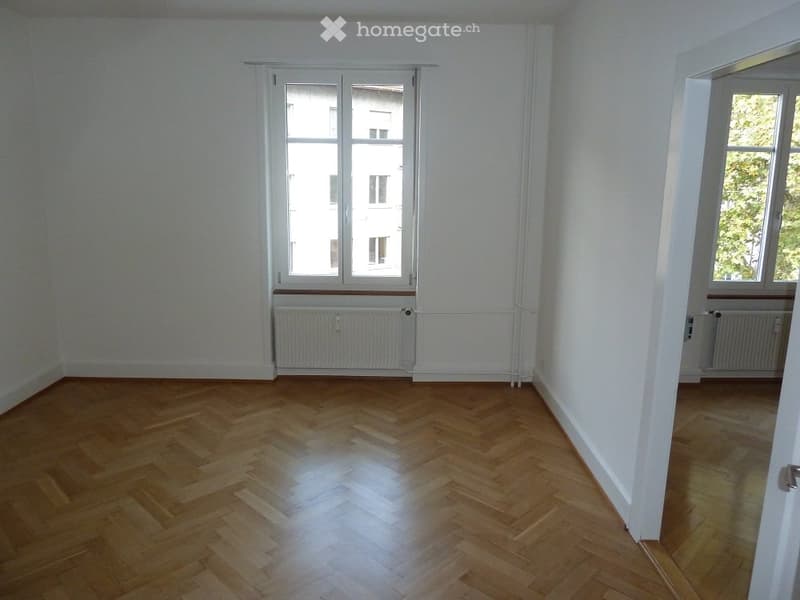 Empty room with wooden herringbone flooring, white walls, large windows, and radiators.