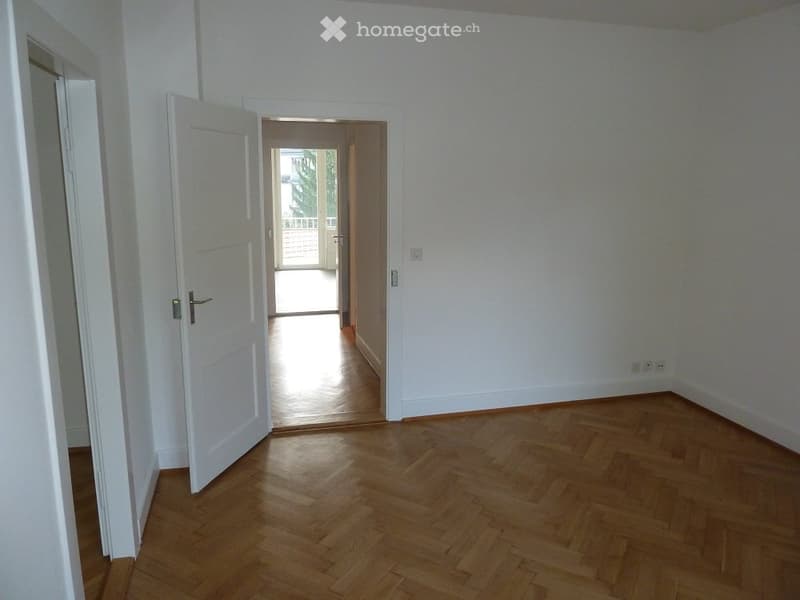 Empty room with wooden flooring and white walls, featuring an open doorway leading to another room and a balcony area visible in the background.