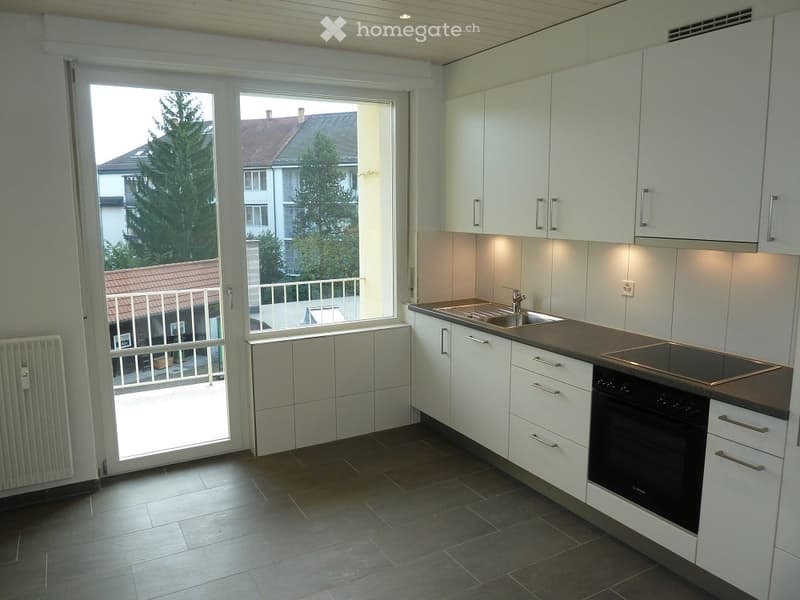 Modern kitchen with white cabinets, dark countertops, and a built-in oven. Large windows and a balcony door provide natural light and access to the outdoor area.