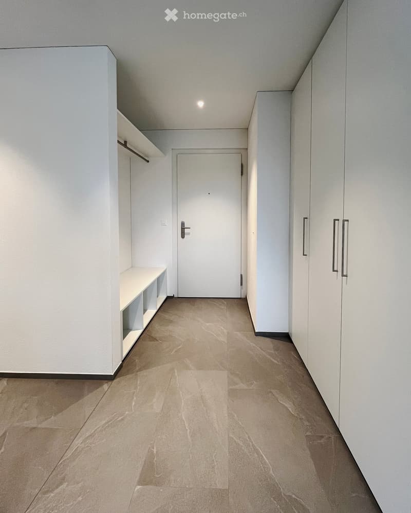 Modern, minimalist hallway with white walls, beige tiled floor, built-in shelving unit, and a closed door at the end.