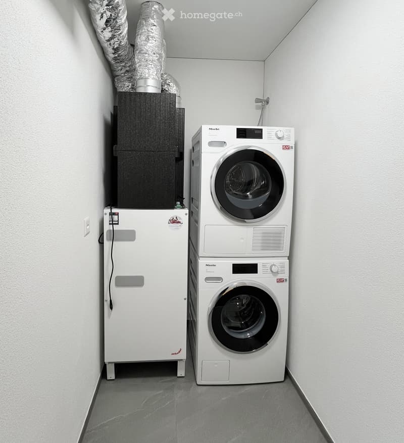 A small, clean utility room with a white washing machine, dryer, and water softener. The walls are white, and the floor is grey.
