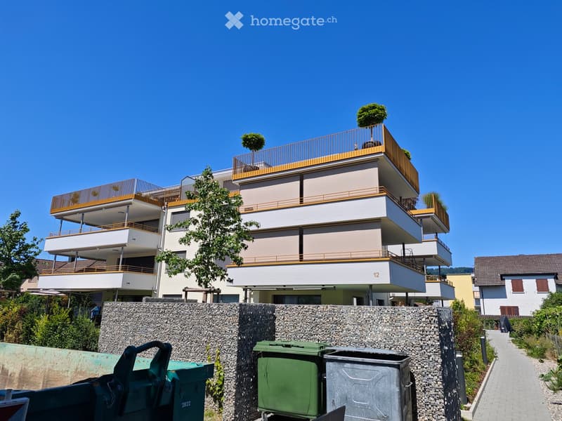Modern multi-story residential building with balconies, beige facade, and golden railings. The building is surrounded by greenery and has a stone wall in the foreground with trash bins.