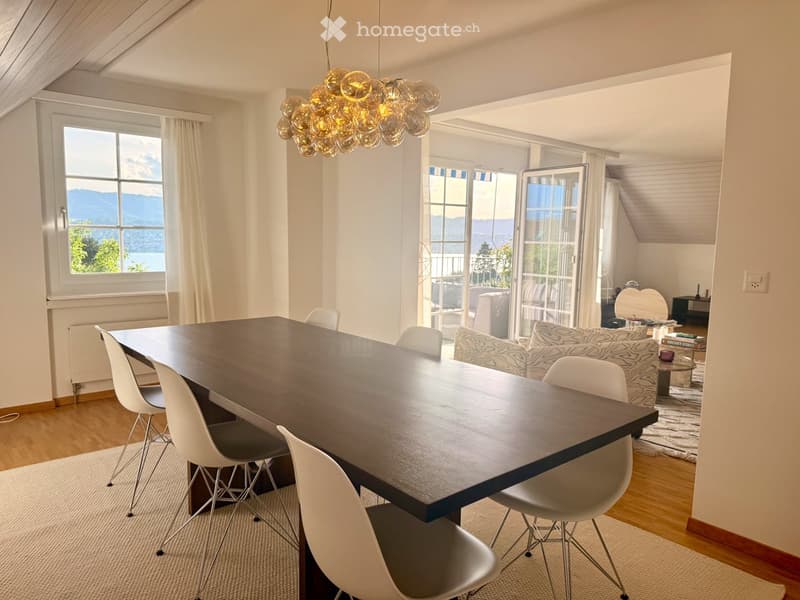 Bright dining area with a large wooden table, white chairs, and a modern chandelier. Adjacent living area features a patterned sofa, coffee table, and scenic mountain view through large windows and sliding doors.