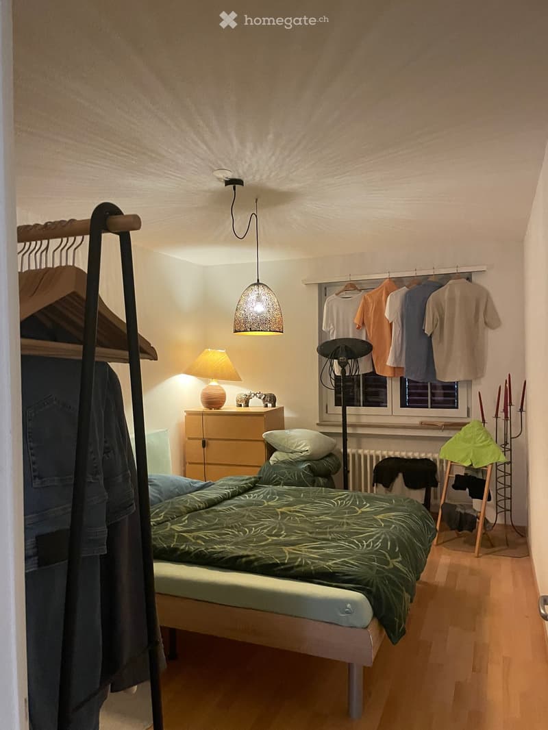 Cozy bedroom with a single bed, nightstand, hanging clothes, and a small wardrobe. The room features wooden flooring and warm lighting.