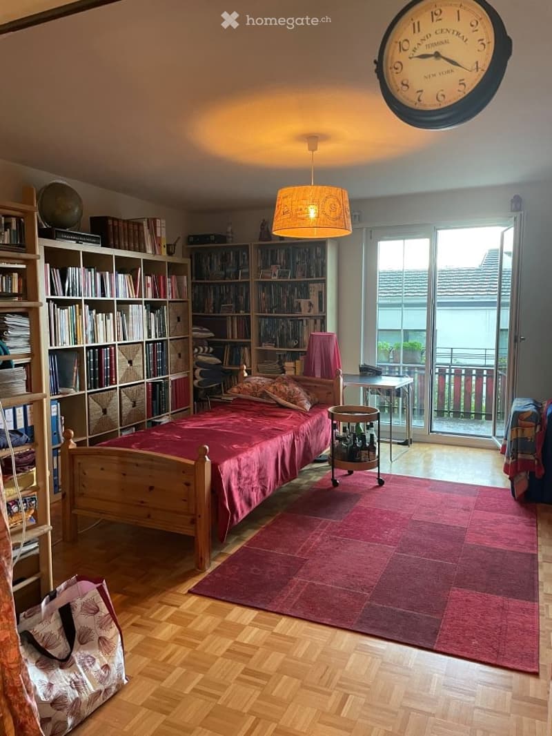 Cozy bedroom with wooden bed, red bedding, bookshelf, small table, and large wall clock.