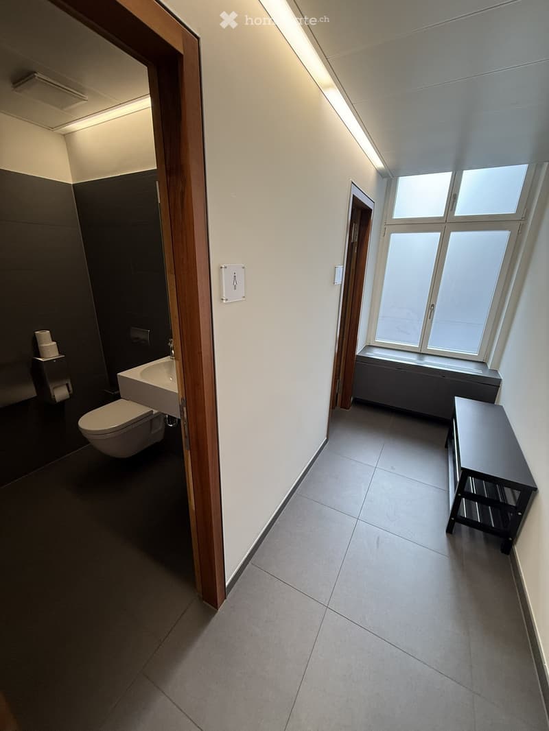 Modern bathroom with a toilet, sink, and a small table. The room has tiled floors and white walls, with a window providing natural light.